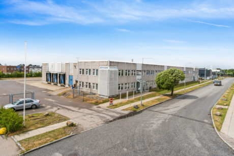 En stor, grå industriel lagerbygning med læsseramper, omgivet af en stort set tom parkeringsplads og en stille gade under en blå himmel.