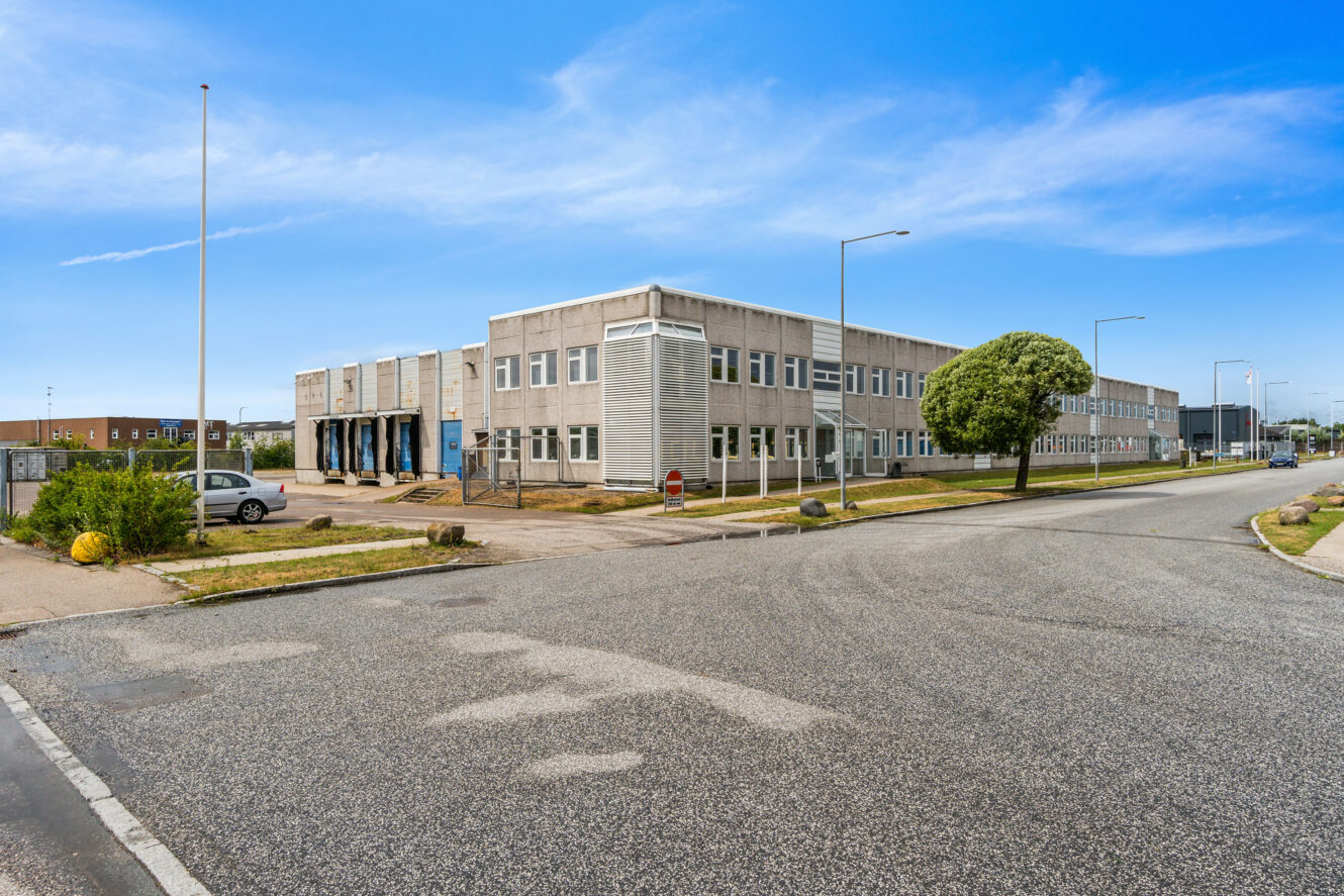 En grå industribygning i to etager ligger på en hjørnegrund med tomme parkeringspladser og et træ i nærheden under en klar blå himmel.