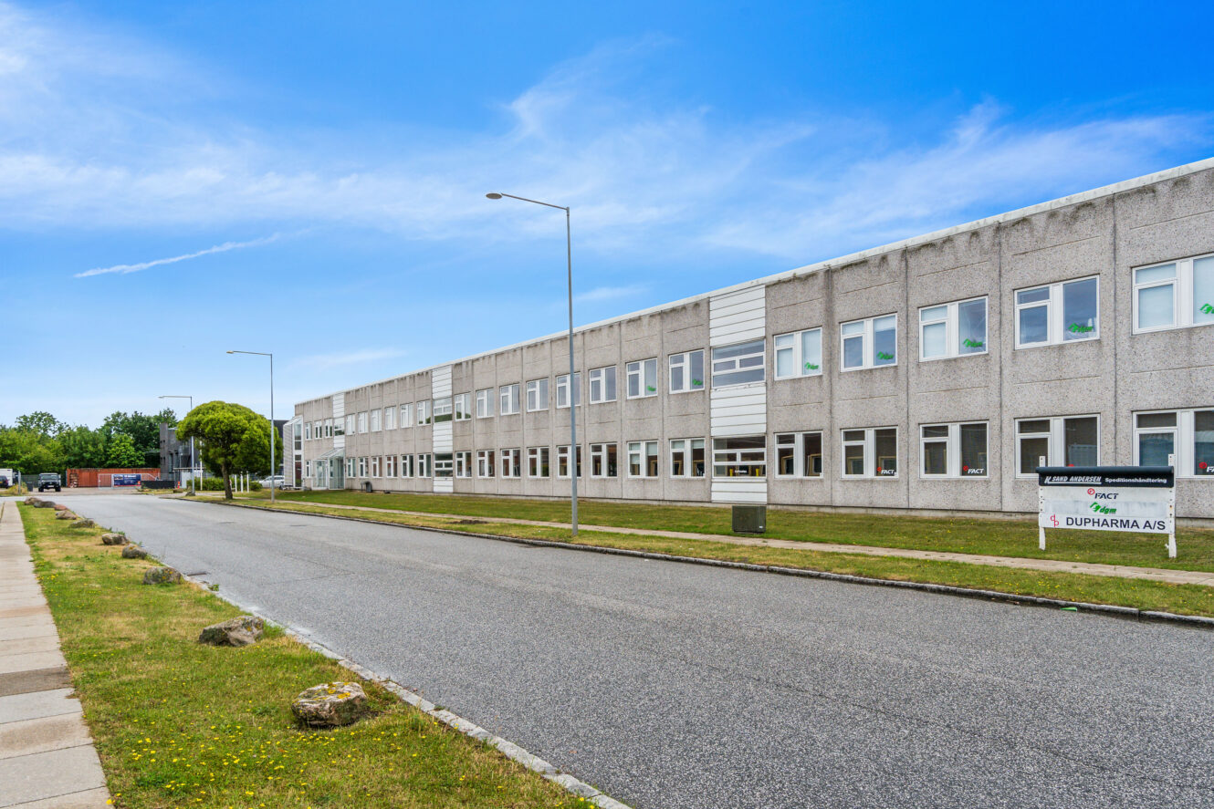 En bred, toetagers grå forretningsbygning med mange vinduer og et skilt foran, der støder op til en stille, tom gade under en klar blå himmel.