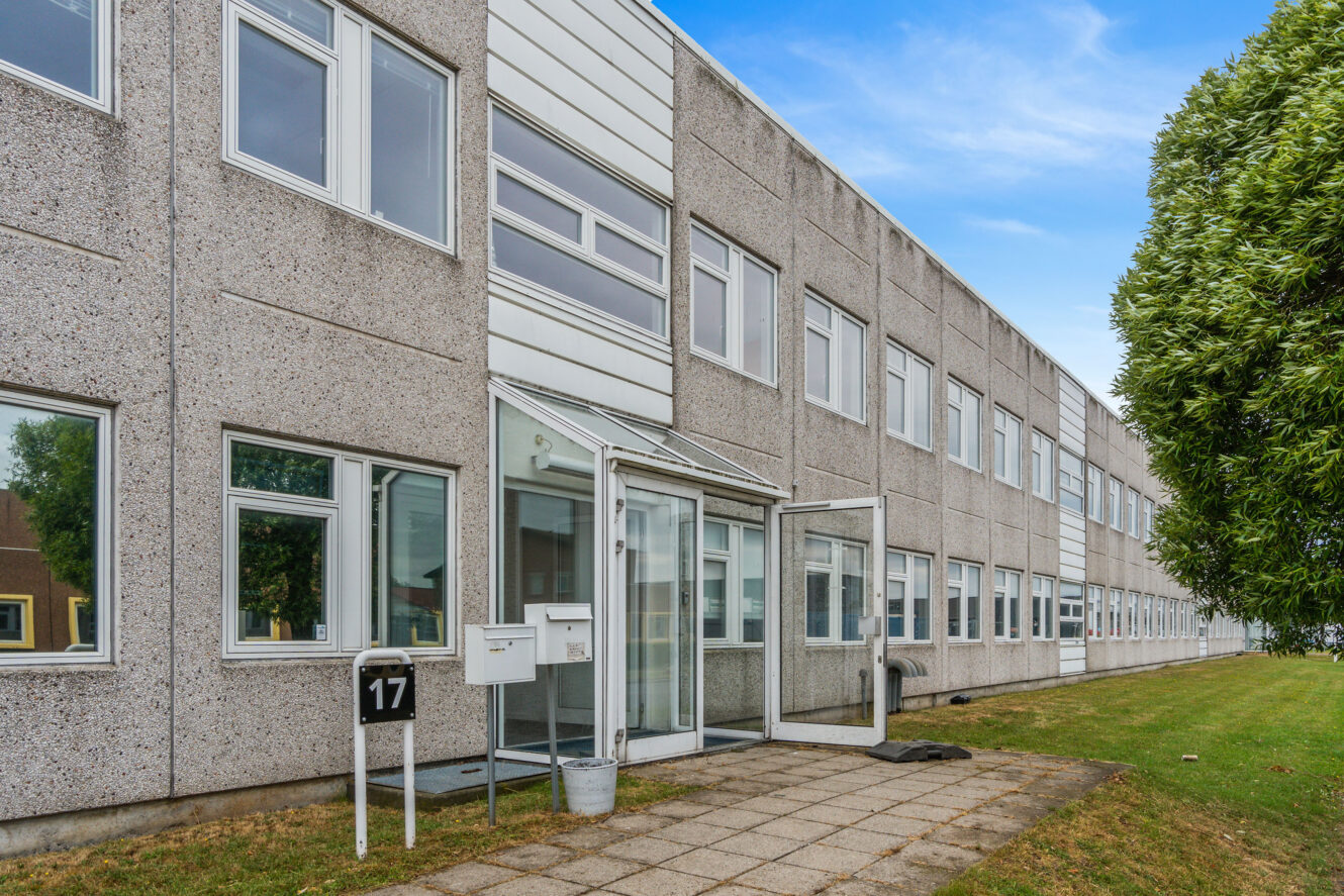En toetagers grå kontorbygning med flere vinduer, en indgangsdør af glas, postkasser mærket 17 og en asfalteret gangsti ved siden af et græsareal og et træ.