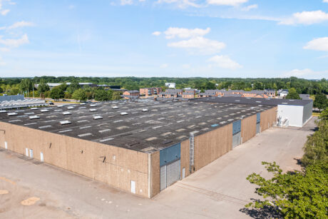 Et stort industrilager med fladt tag og flere ovenlysvinduer ligger i et industriområde med træer og beboelsesbygninger i baggrunden.