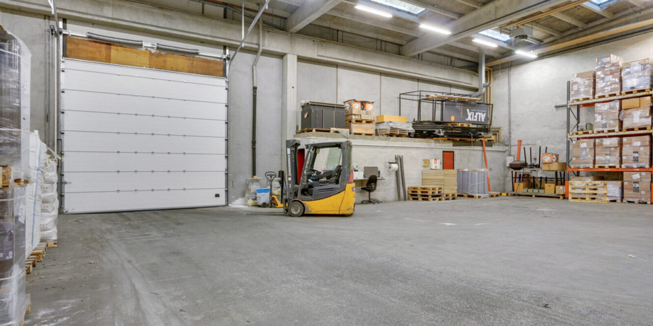 En gul gaffeltruck står i en rummelig lagerhal med højt til loftet, hylder fyldt med kasser og en stor lukket port til læsserampen.