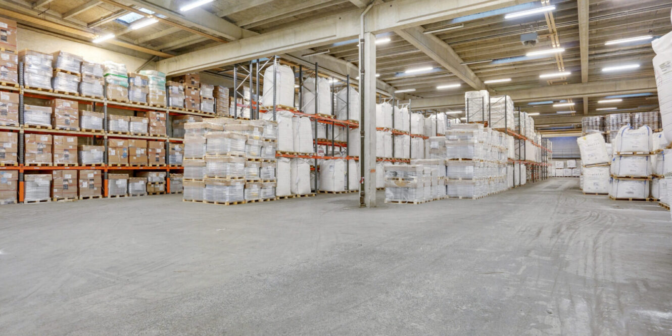 Stort lagerinteriør med høje hylder og paller stablet med kasser og poser, organiseret i rækker under kraftig industriel belysning.