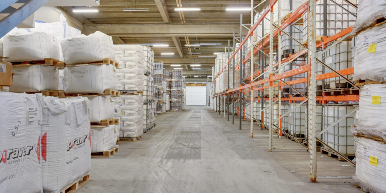 Industrielt lagerinteriør med stablede hvide bulkposer på paller til venstre og metalreoler med store containere til højre, under fluorescerende belysning.