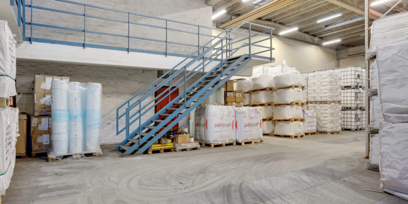 Industrielt lagerinteriør med stablede paller med varer, store sække og ruller med materiale. En metaltrappe fører op til en øvre mezzanin. Området er godt oplyst med loftsbelysning.