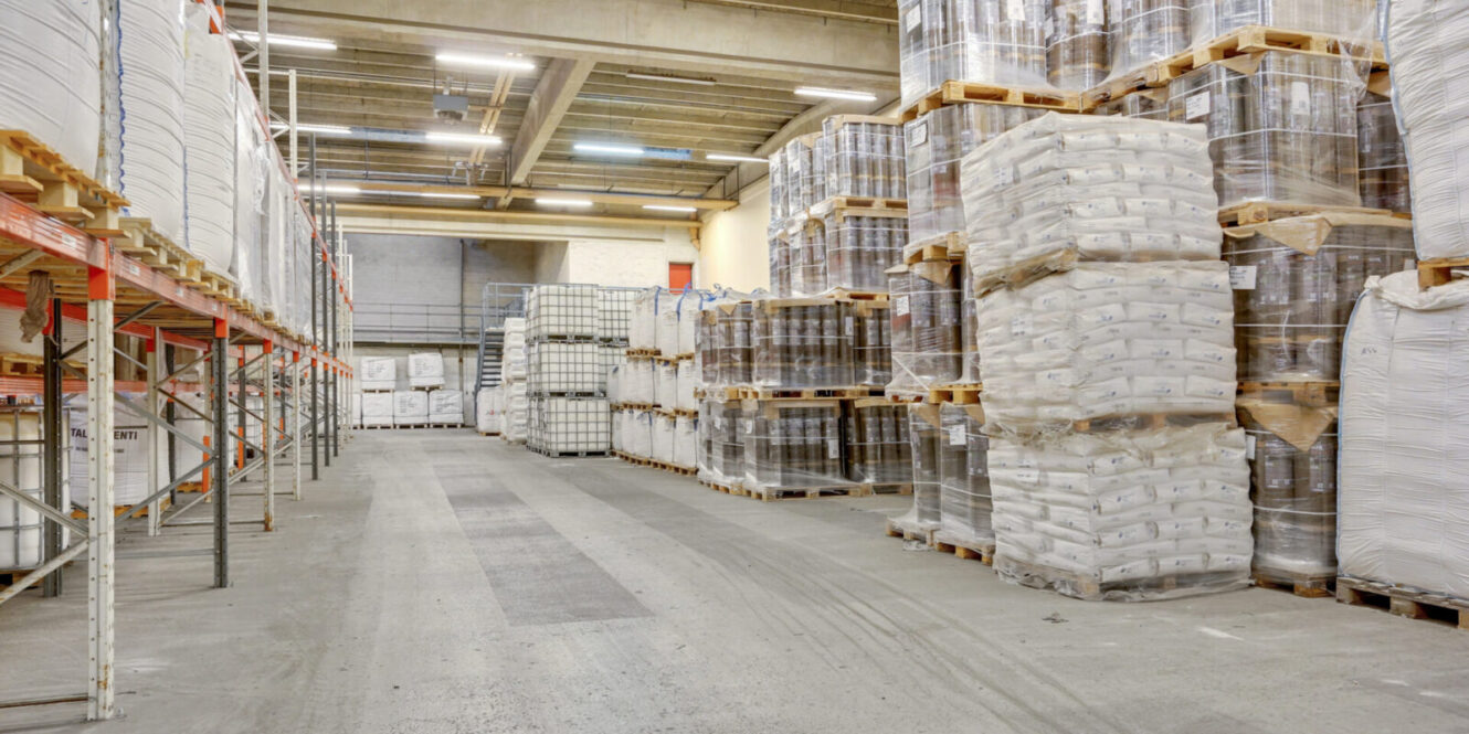 Et lagerinteriør med paller stablet højt med indpakkede poser og beholdere, opstillet langs brede reoler og betongulve.