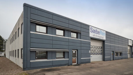En stor, grå industribygning med flere vinduer og et skilt over en garageport, hvor der står "SæbeRRen - alt til rengøring".