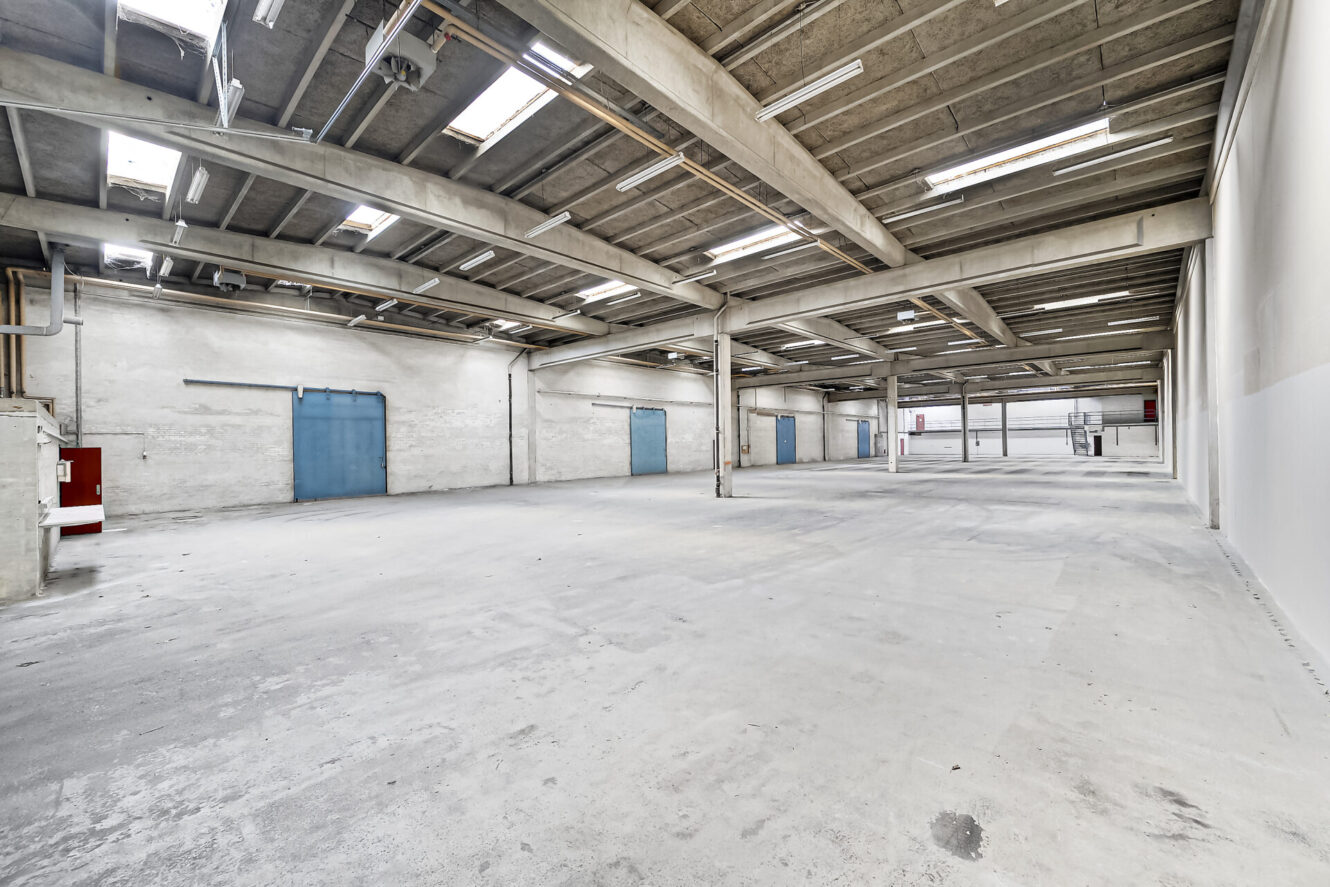 Stort, tomt industrilager med betongulv, synlige bjælker, fluorescerende belysning og blå metaldøre langs sidevæggene.