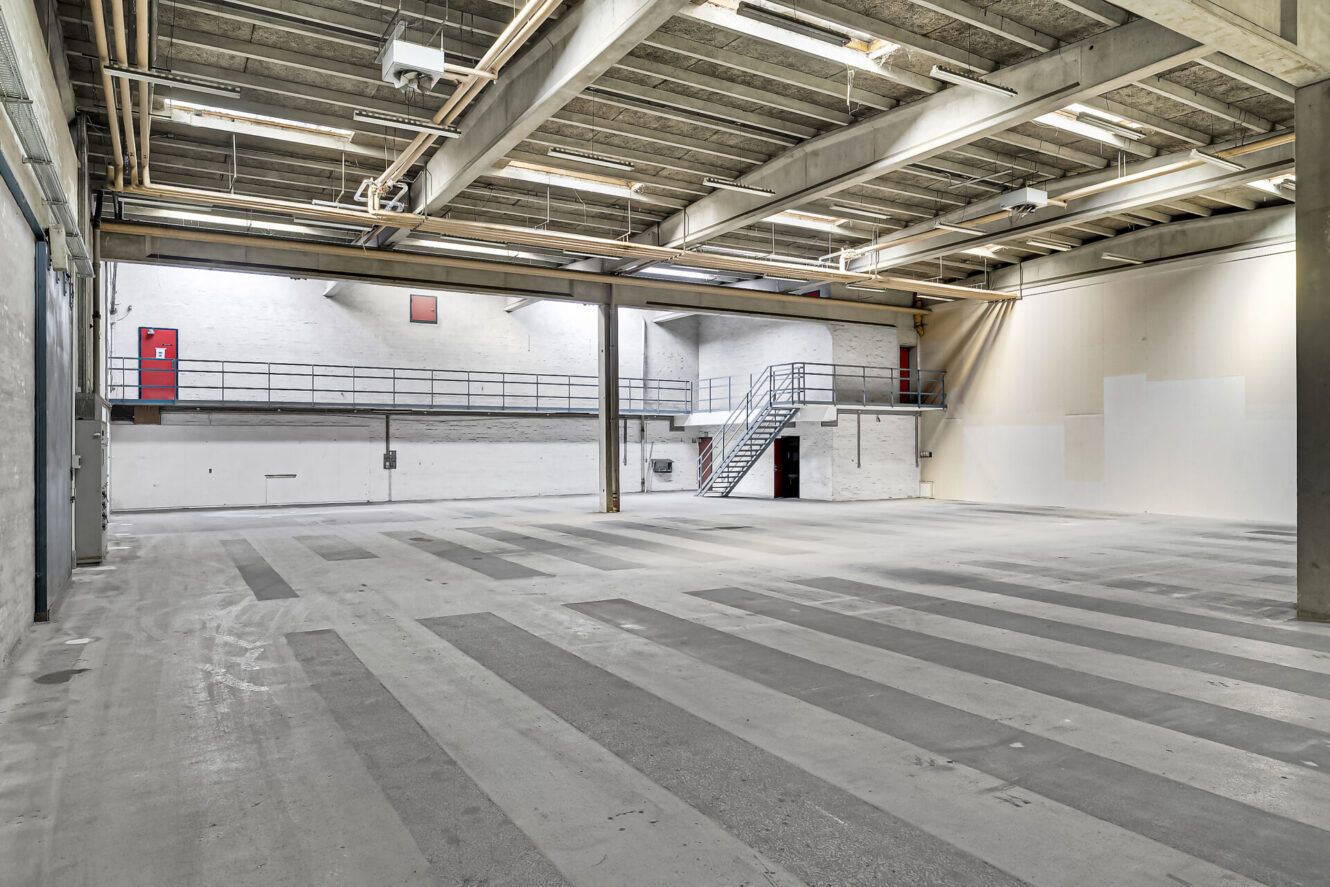 Rummeligt, tomt lagerinteriør med betongulv, synlige bjælker, højt til loftet, mezzanin-niveau, metaltrapper og hvidmalede vægge.