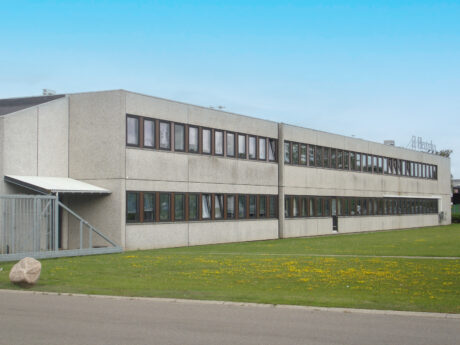 En rektangulær kontorbygning i to etager med mange vinduer, et fladt tag og et ydre af beton, der ligger på en græsplæne under en klar blå himmel.