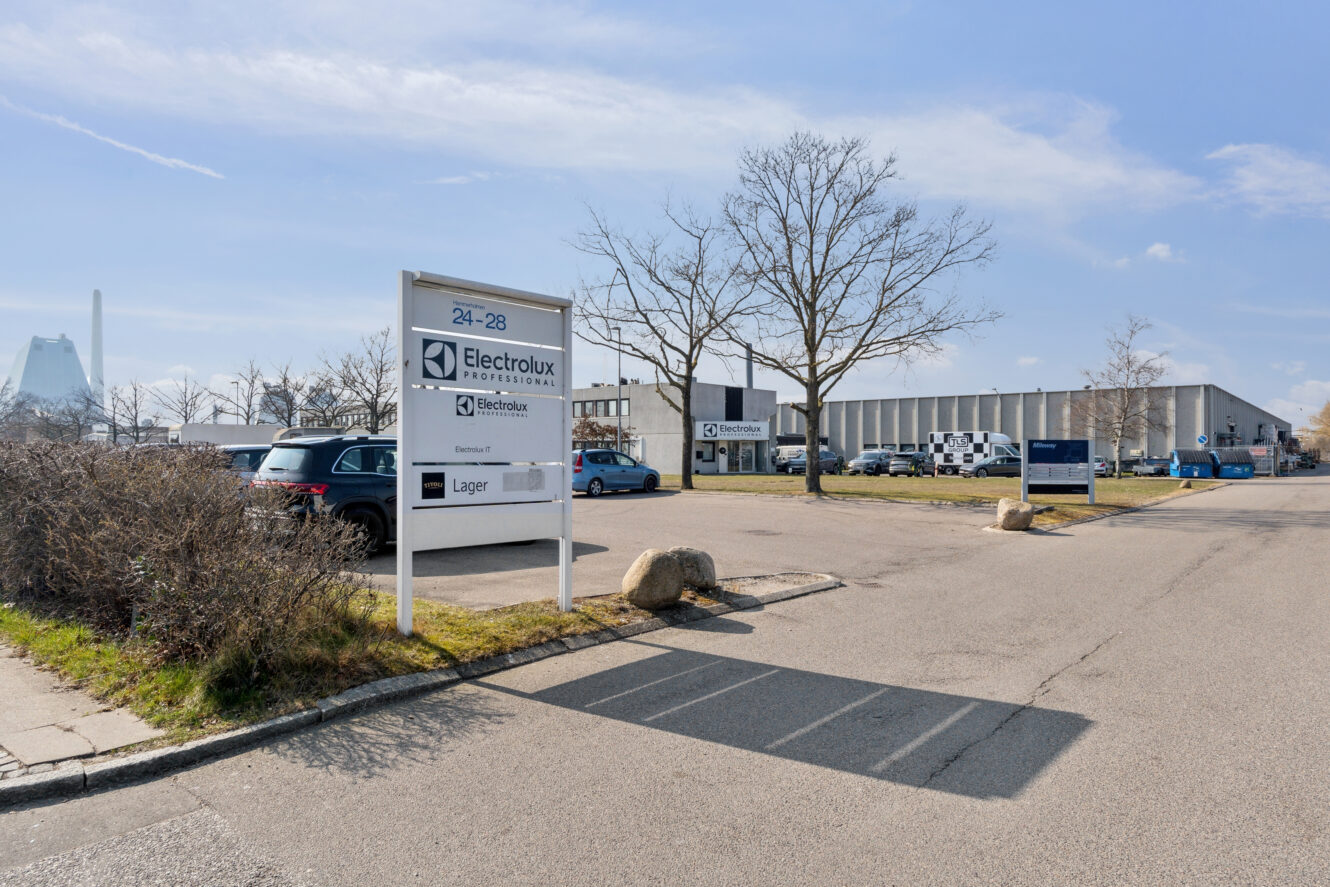 En parkeringsplads med flere biler foran en industribygning med Electrolux Professional-skilte og løvløse træer langs indkørslen.