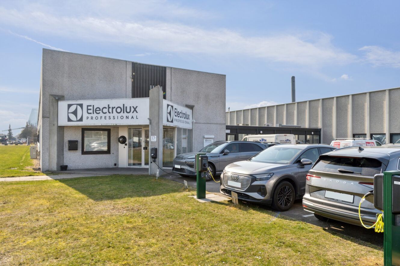 Udvendigt på en Electrolux Professional-bygning med flere parkerede biler, herunder en elbil, der er tilsluttet en ladestation i forgrunden.