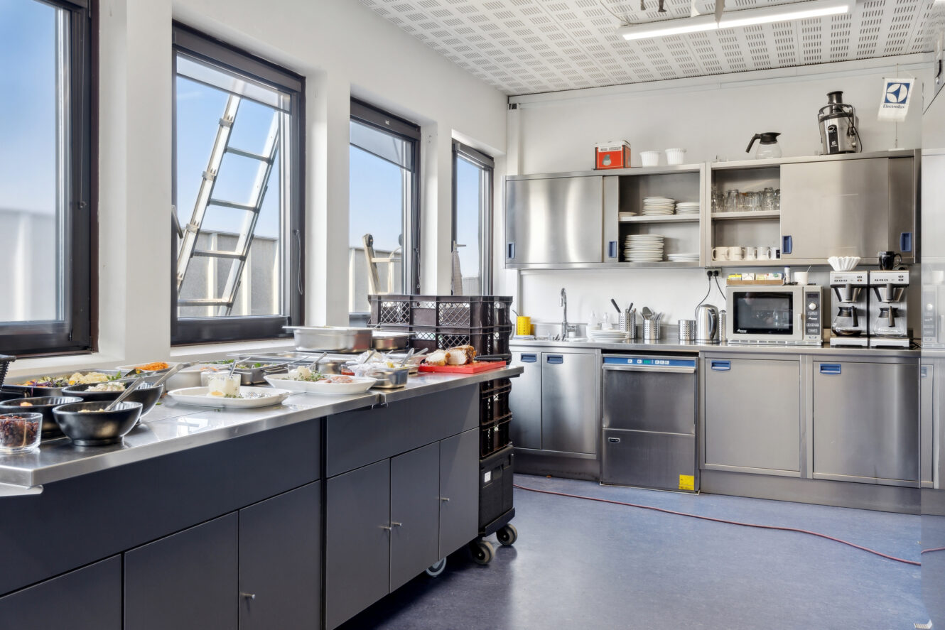 Et moderne storkøkken med apparater i rustfrit stål, tallerkener og mad lagt på en disk, oplyst af naturligt lys fra flere store vinduer.