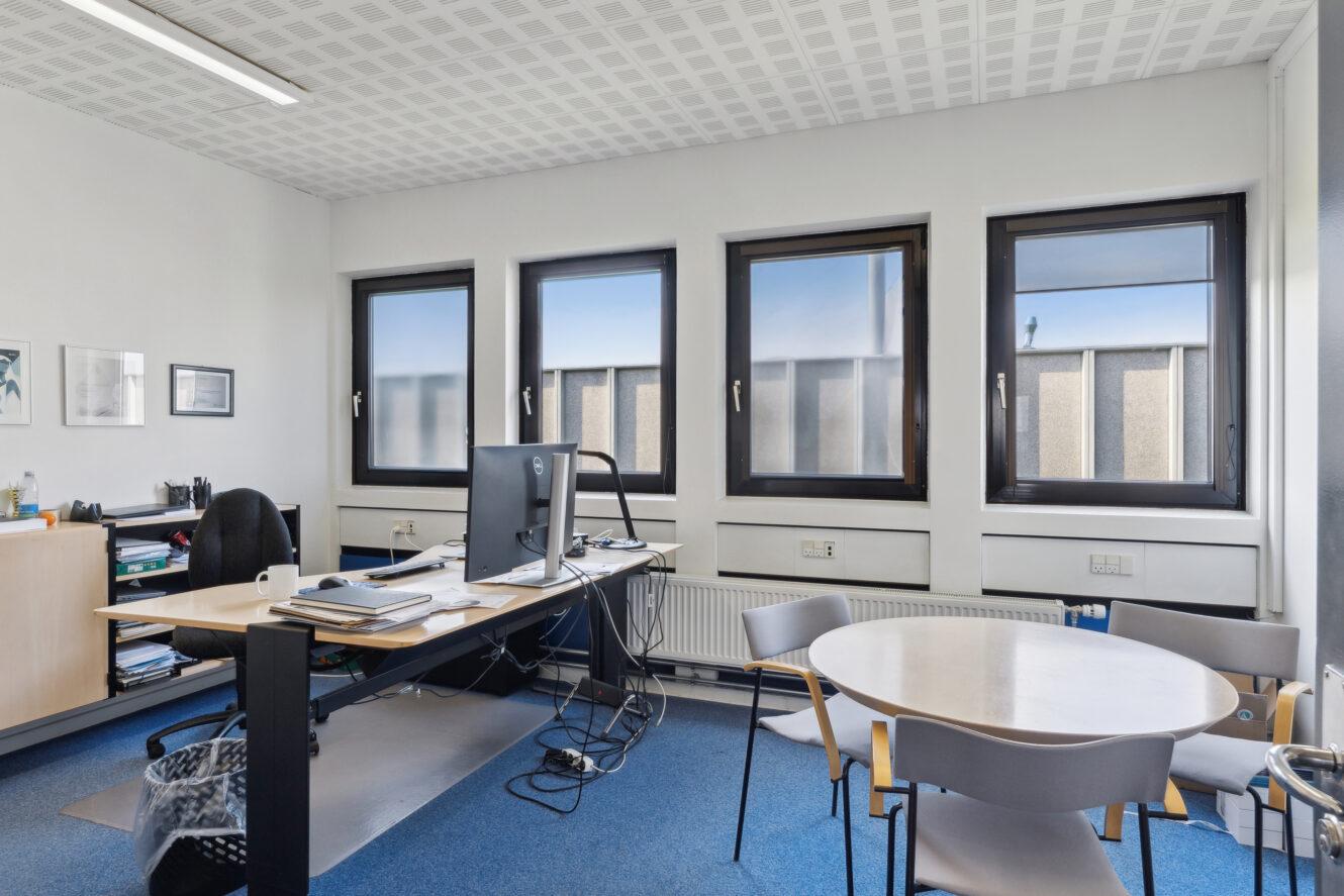 Moderne kontorlokale med skrivebord, computer, kontorstol, hylder og et lille rundt bord med fire stole og tre store vinduer.