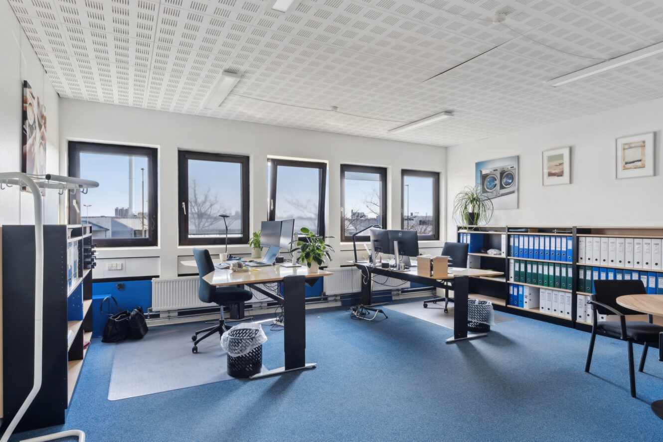 Moderne kontor med to skriveborde, kontorstole, computere, planter, hylder med ringbind og store vinduer, der lukker naturligt lys ind.