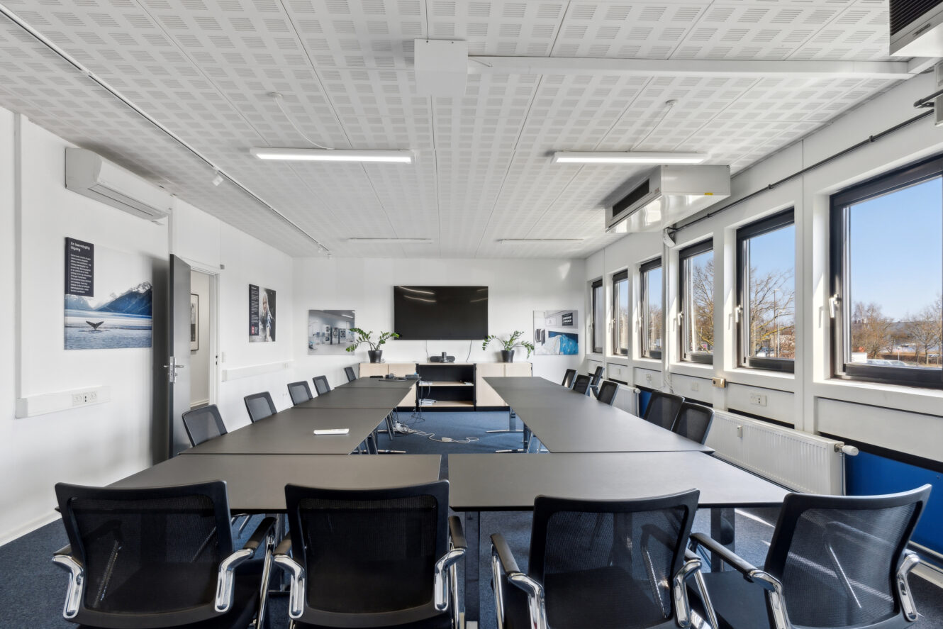 Et moderne konferencelokale med sorte stole, rektangulære borde arrangeret i en U-form, store vinduer, vægkunst og et fladskærms-tv foran.