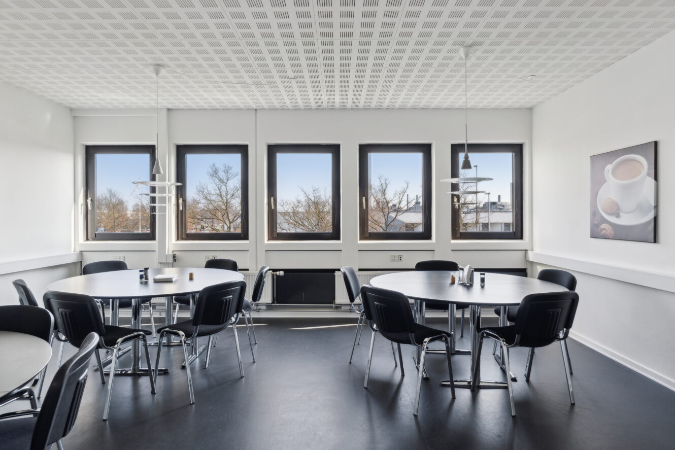 Et lyst, moderne pauserum med fire runde borde, sorte stole, pendellamper og vinduer med udsigt til træer; et billede med kaffetema hænger på væggen.