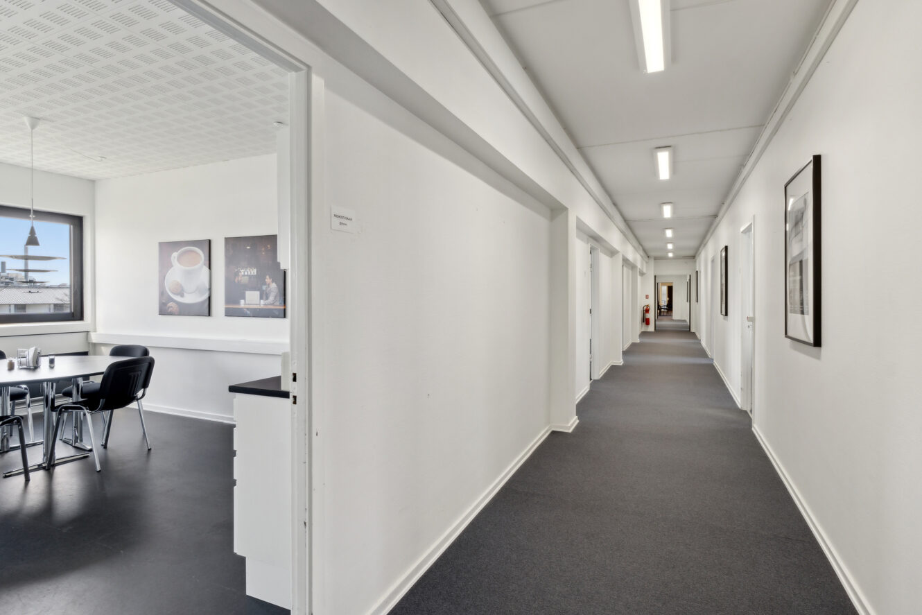 En ren, veloplyst kontorgang med hvide vægge og flere døre, der støder op til et lille mødelokale med et bord, stole og indrammede kunstværker med kaffetema.