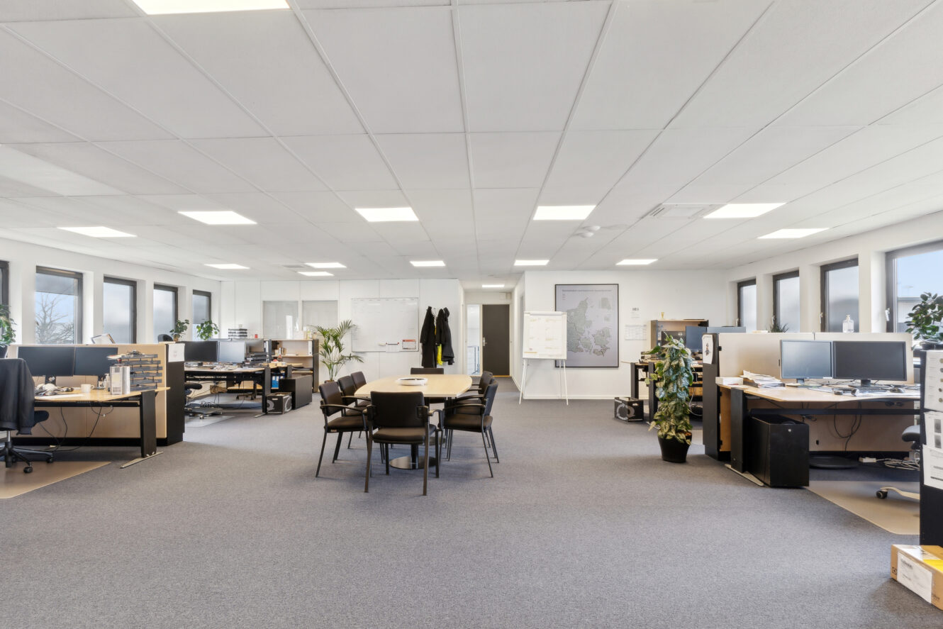 Moderne storrumskontor med flere skriveborde, computere, kontorstole, planter, et centralt mødebord, whiteboards og store vinduer, der giver naturligt lys.