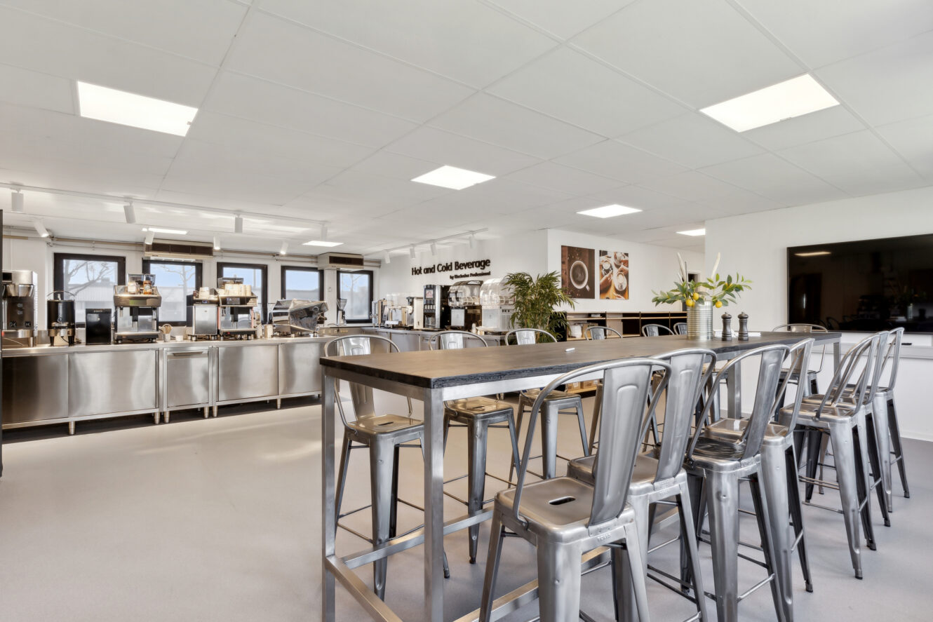 Et moderne cafeteria med metalborde og -stole, skranker i rustfrit stål, drikkevareautomater og kraftig ovenlys.