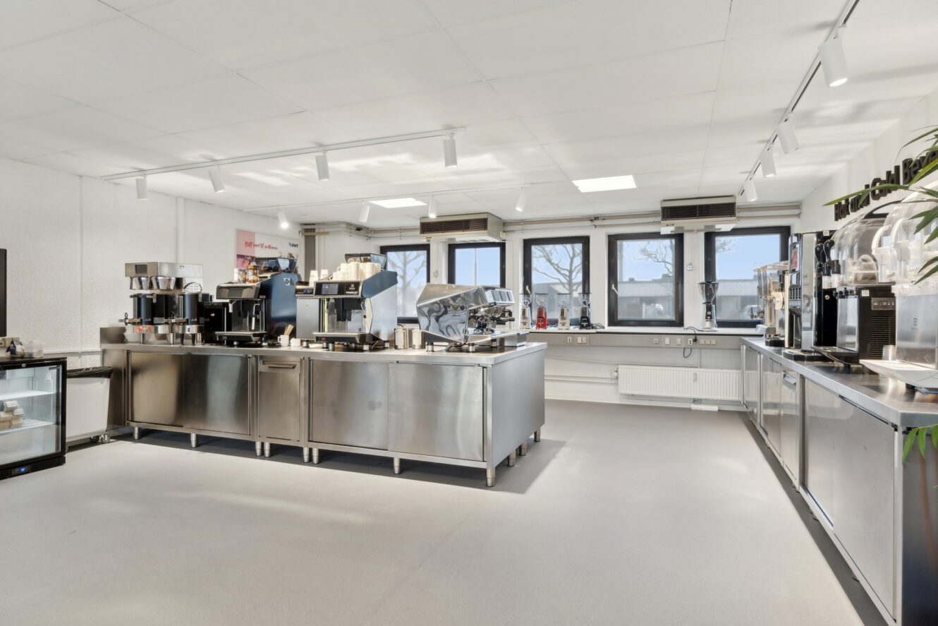 Moderne storkøkken med apparater i rustfrit stål, bordudstyr, kaffemaskiner og store vinduer, der lukker naturligt lys ind.