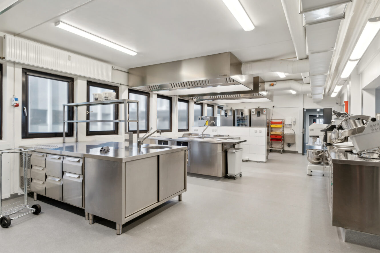 Et moderne, rummeligt storkøkken med apparater i rustfrit stål, arbejdsborde, hylder og kraftig belysning.