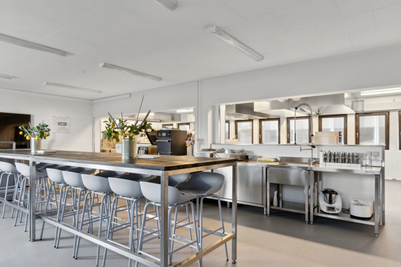 Moderne storkøkken med hvidevarer i rustfrit stål, lang træø med barstole og blomsterarrangementer på disken. Rummet er lyst med ovenlys.