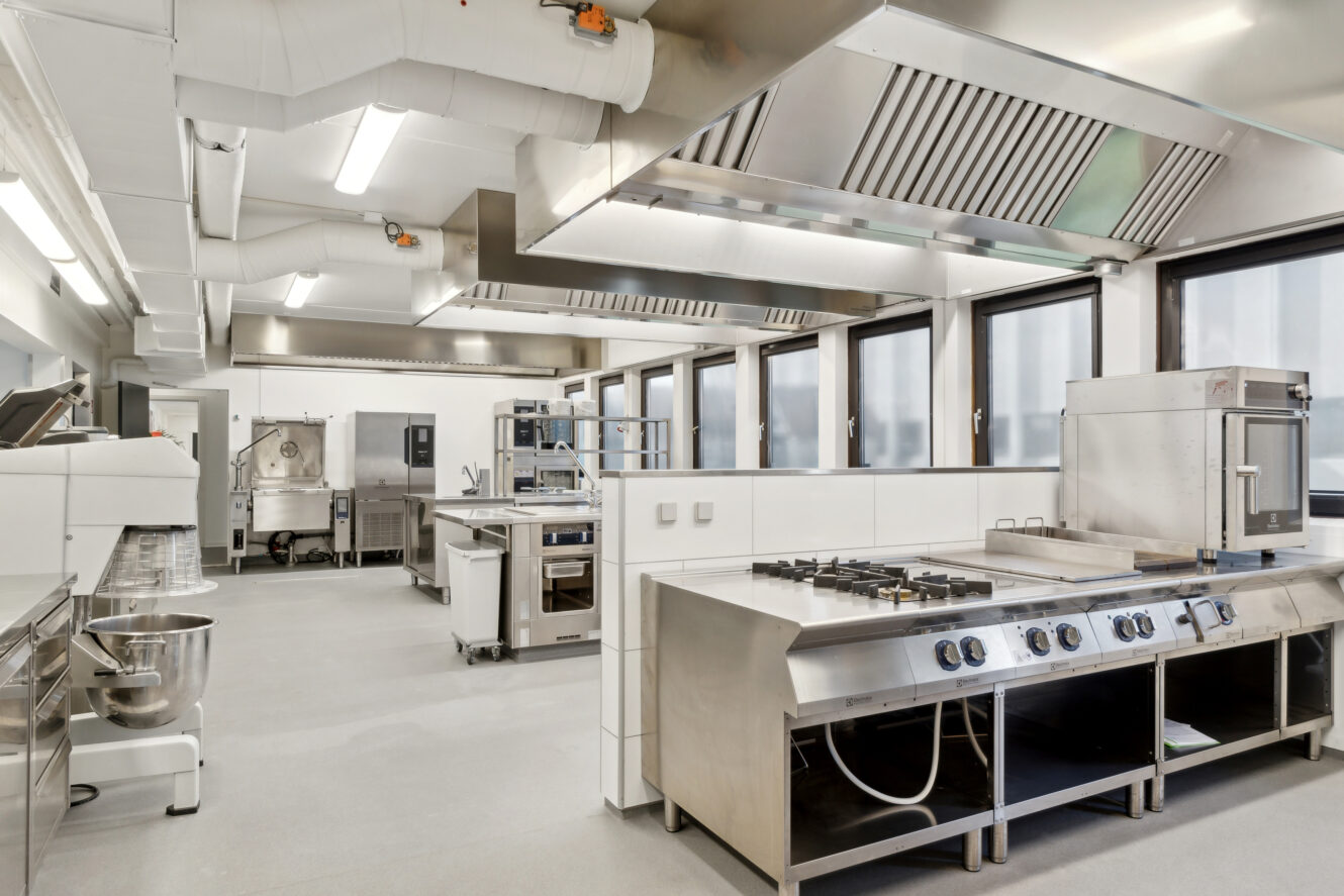 Et moderne storkøkken med apparater i rustfrit stål, gaskomfurer, ventilationshætter og rene bordplader, godt oplyst af loftslamper og store vinduer.