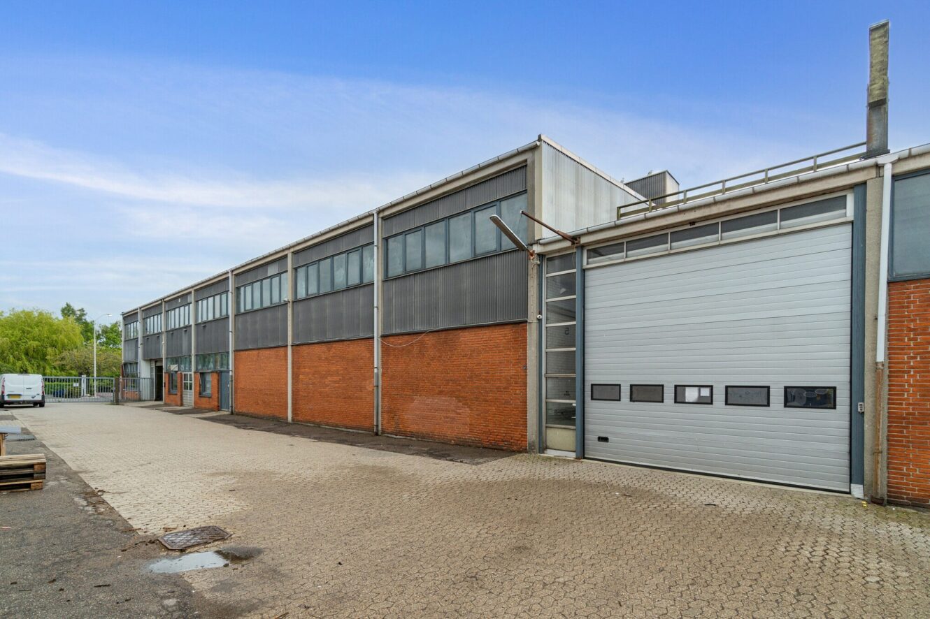 Stor industribygning med undervægge af røde mursten, overvægge af metal og en stor hvid rulleport under en blå himmel.