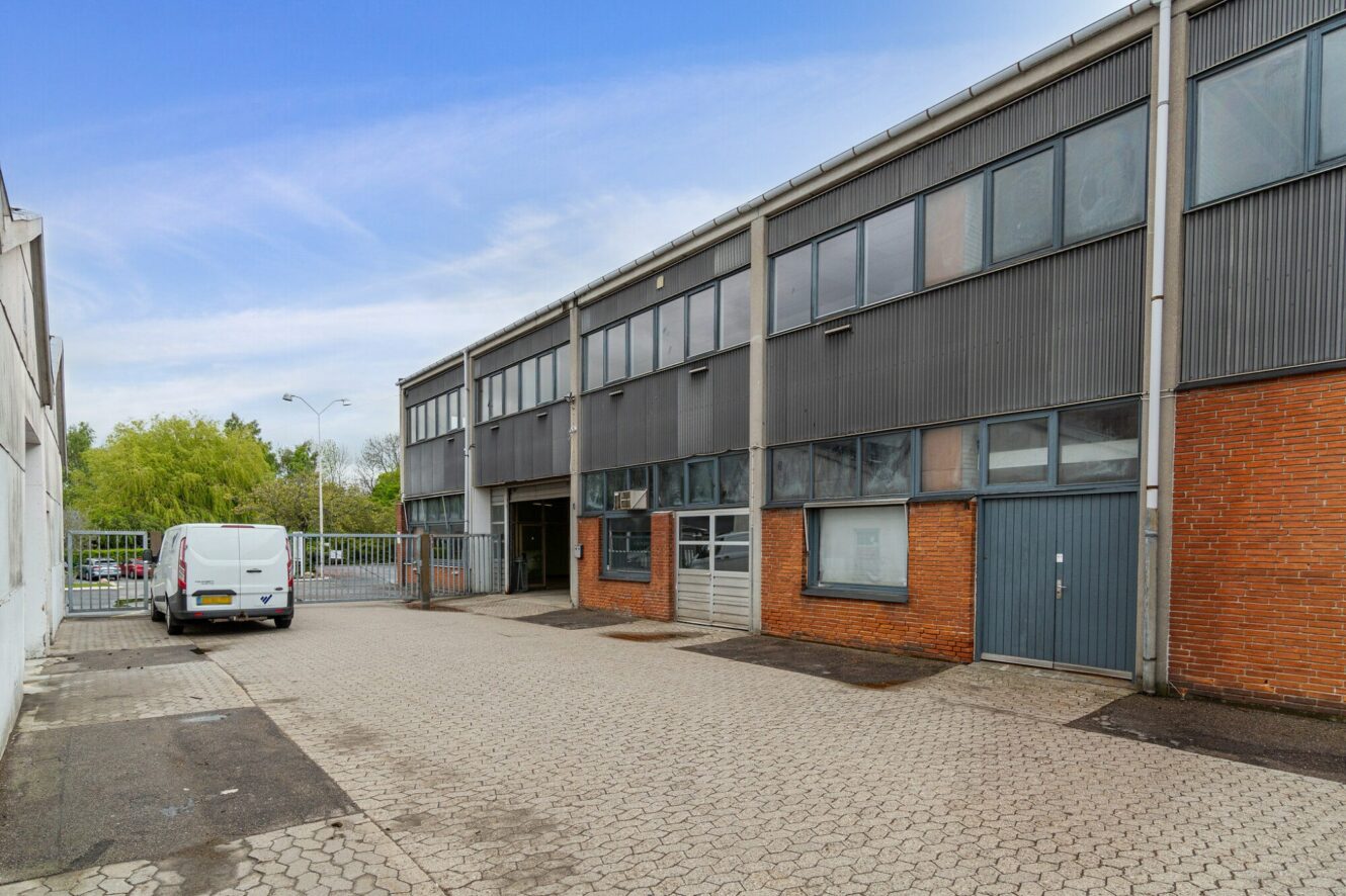 En hvid varevogn er parkeret uden for en industriel lagerbygning med store vinduer, mursten og metalbeklædning og en indhegnet indgang på en asfalteret grund.