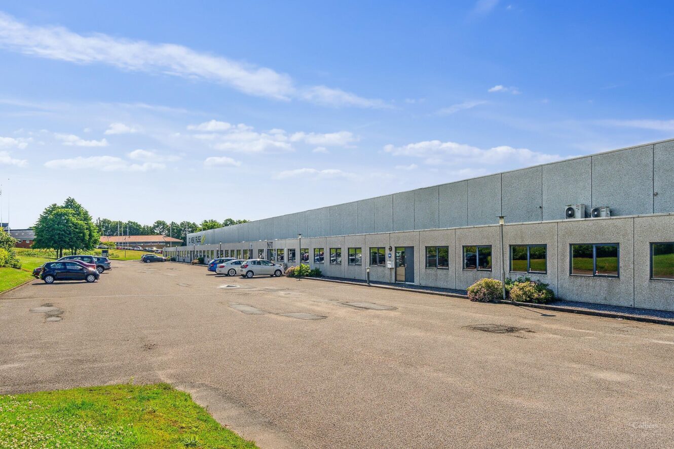 En stor erhvervsbygning i én etage med mange vinduer og en stort set tom parkeringsplads på en solrig dag.
