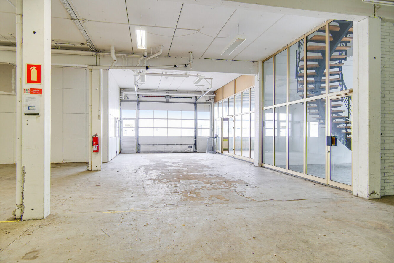 Rummeligt, tomt industriinteriør med betongulv, store vinduer, kontorområde med glasvægge og en trappe, der fører op til en øvre etage.