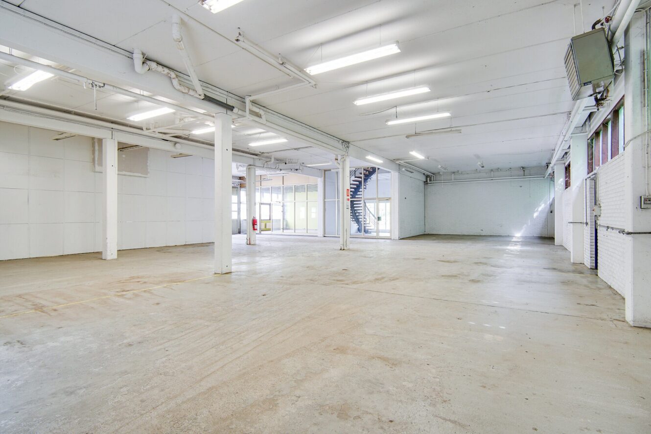Stort, tomt industrilager med hvide vægge, betongulve, synlige rør, lysstofrør og en trappe, der fører op til en mezzanin. Naturligt lys kommer ind gennem vinduer og glasdøre.