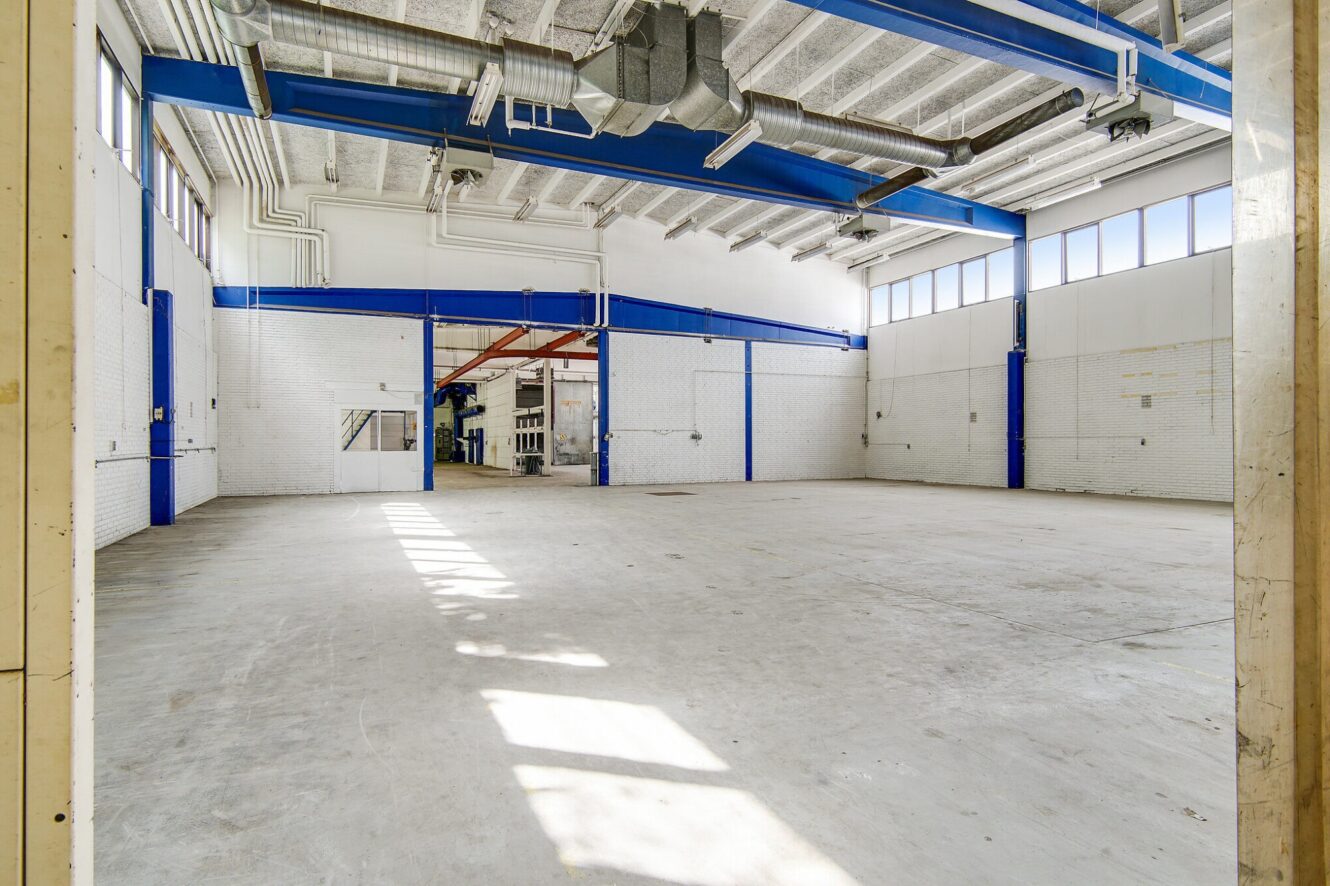 Stort, tomt industrilager med hvide vægge, betongulv, højt til loftet, synlige kanaler og blå stålbjælker. Sollyset strømmer ind fra høje vinduer langs den ene væg.
