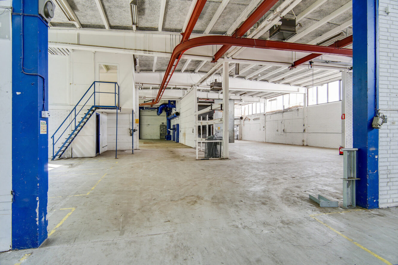 Stort tomt industrilager med højt til loftet, hvide murstensvægge, blå stålbjælker, store vinduer og en metaltrappe, der fører op til en mezzanin.