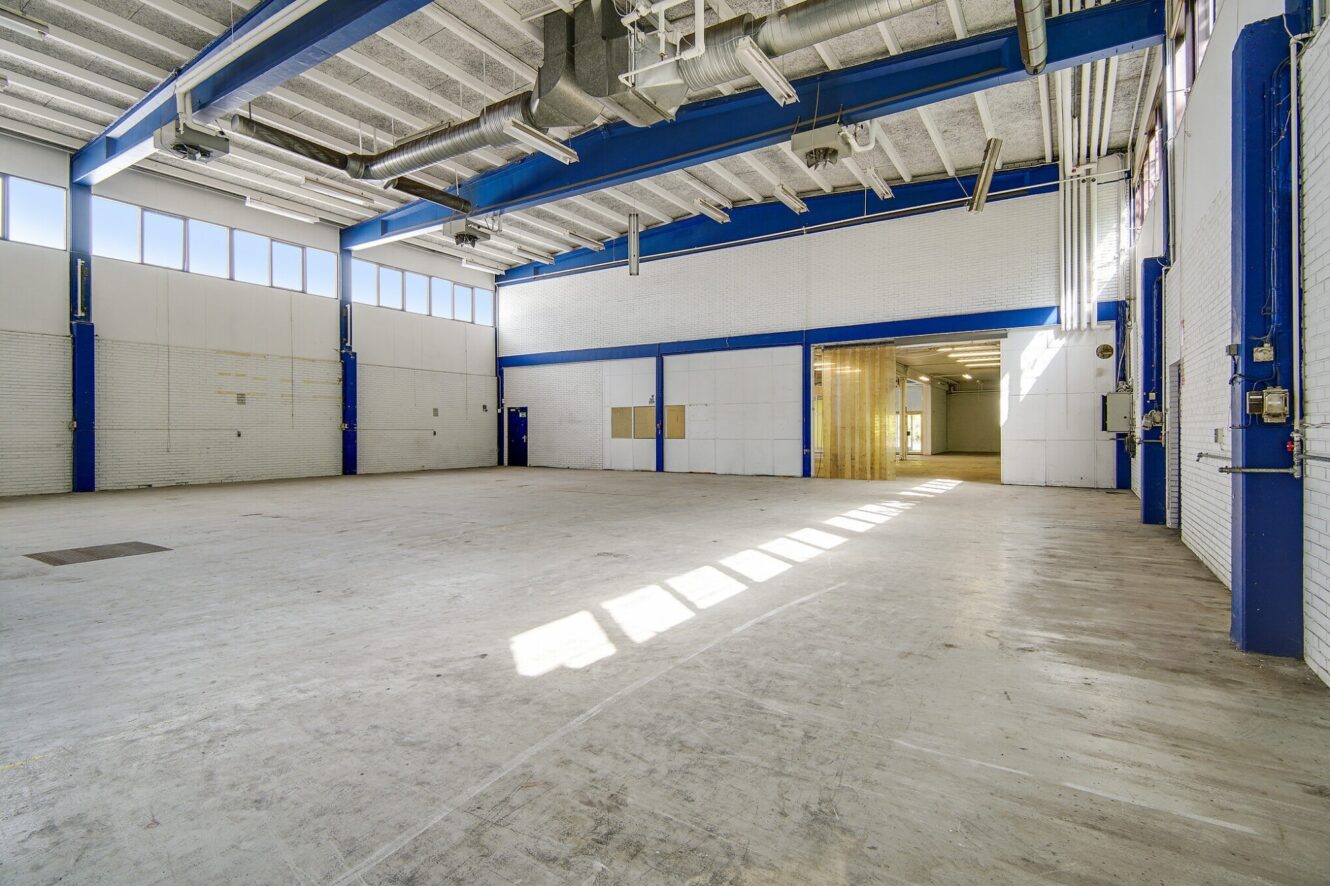 Tomt industrilager med højt til loftet, synlige kanaler, hvide vægge og blå stålbjælker. Sollyset strømmer ind gennem høje vinduer og ned på betongulvet.