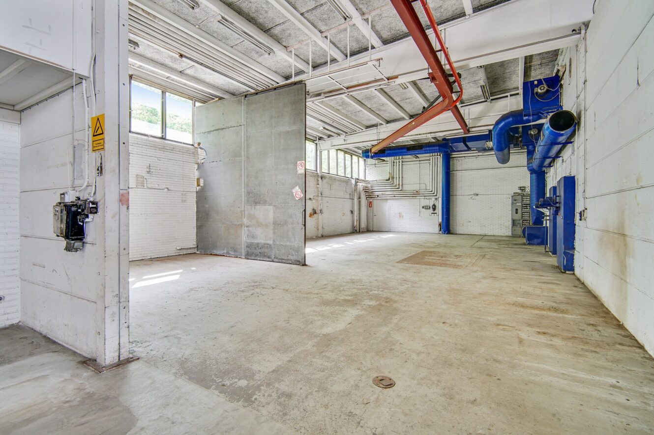 Stort, tomt industrilager med hvide murstensvægge, synlige loftsrør, betongulv og blåt ventilationsudstyr monteret på højre væg. Naturligt lys kommer ind gennem vinduer.