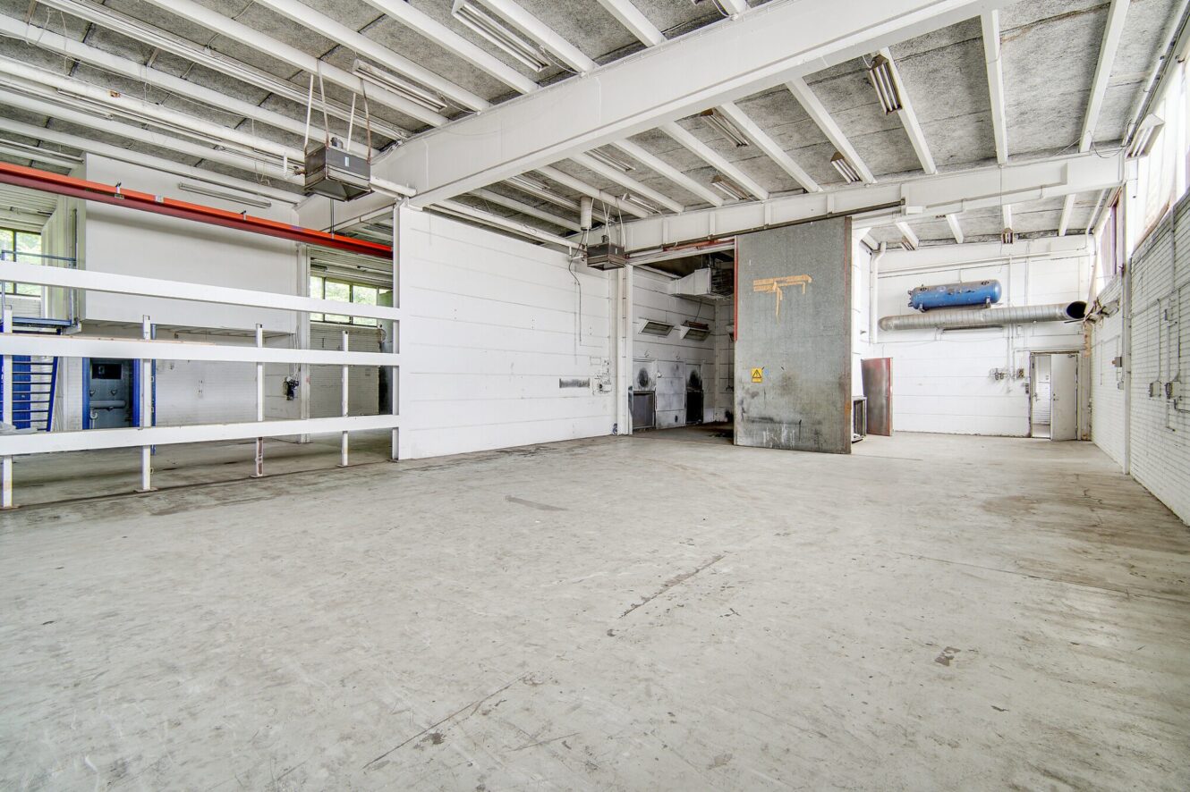 Interiør af et tomt industrilager med hvide vægge, betongulv, synlige loftsbjælker og forskellige rør og inventar.