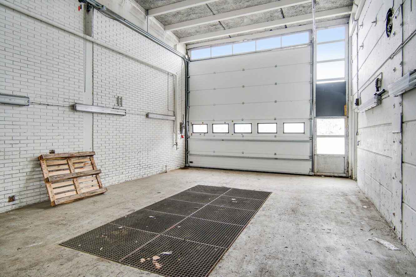 Tom industrigarage med hvide murstensvægge, højt til loftet, en stor hoveddør med vinduer, metalriste på gulvet og en træpalle, der læner sig op ad væggen.