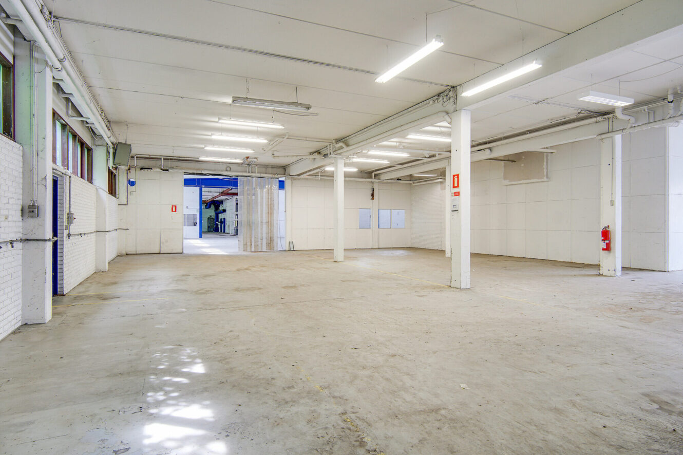 Stort tomt industrilager med hvide vægge, stort åbent gulv, synlige rør i loftet, lysstofrør og en åben garageport, der fører ud.