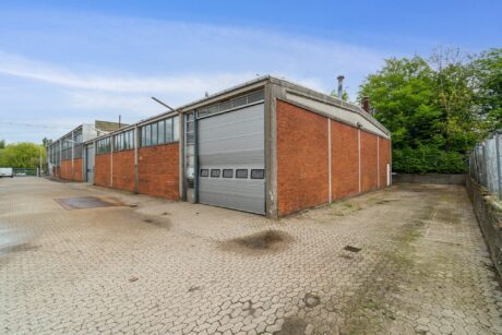 Et stort industrielt lager i mursten med en grå rulleport, der ligger på en asfalteret grund med træer og et metalhegn i baggrunden.