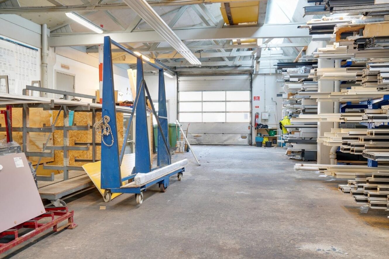 Industrielt lagerinteriør med metalhylder med forskellige materialer, store blå vogne med glasruder og udstyr, der er organiseret langs væggene under et skylit-loft.