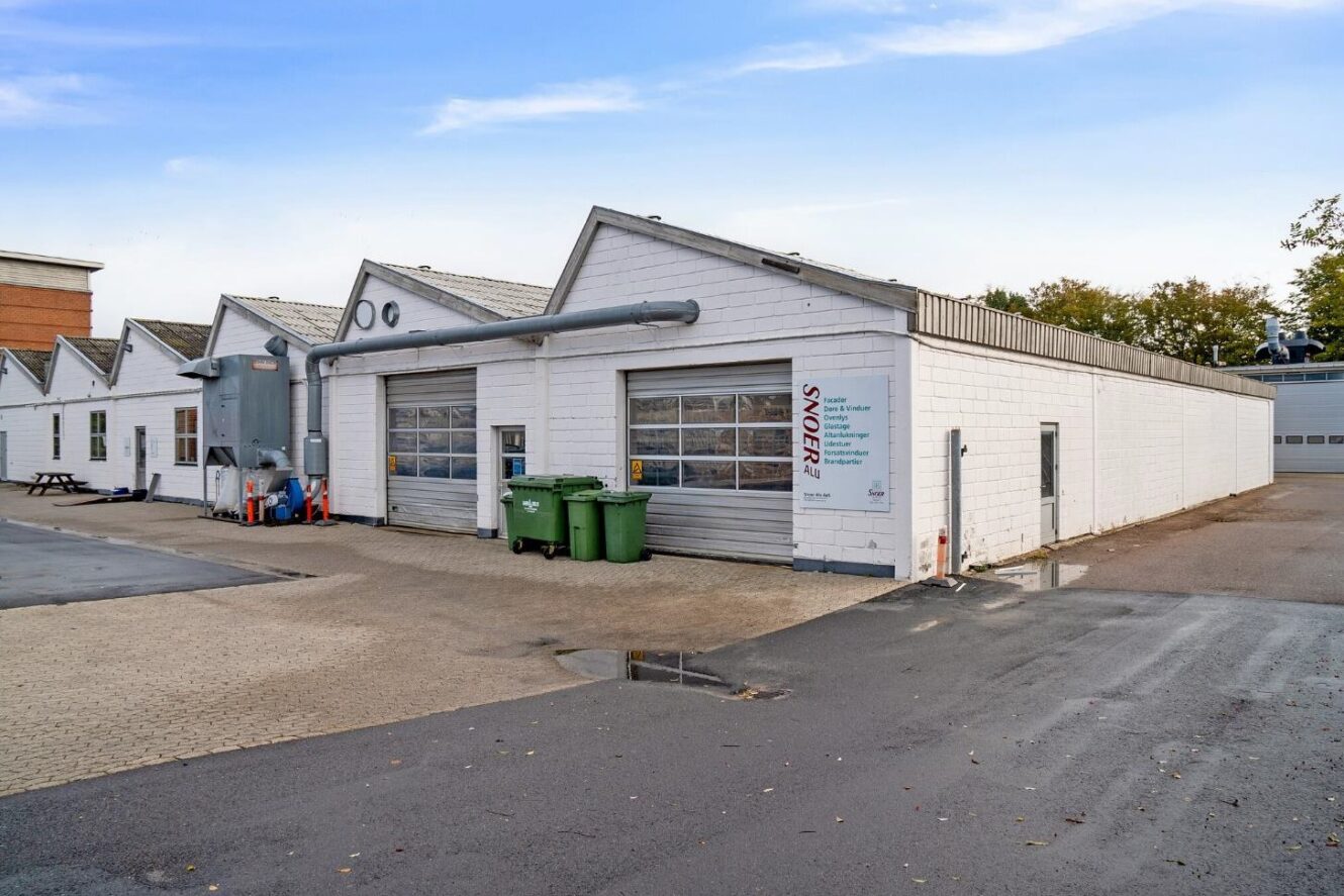 En række hvide industribygninger med store garageporte, grønne skraldespande og udendørs rør ligger på en asfalteret grund under en delvis overskyet himmel.