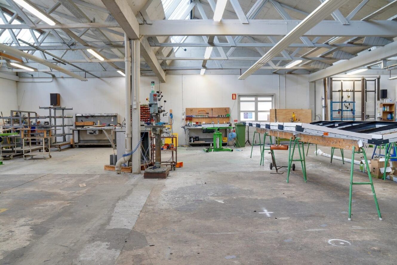 Industrielt værksted med arbejdsbænke, maskiner, værktøj og udstyr under et højt loft. Rummet er organiseret og godt oplyst, og de forskellige materialer og arbejdsstationer er synlige.