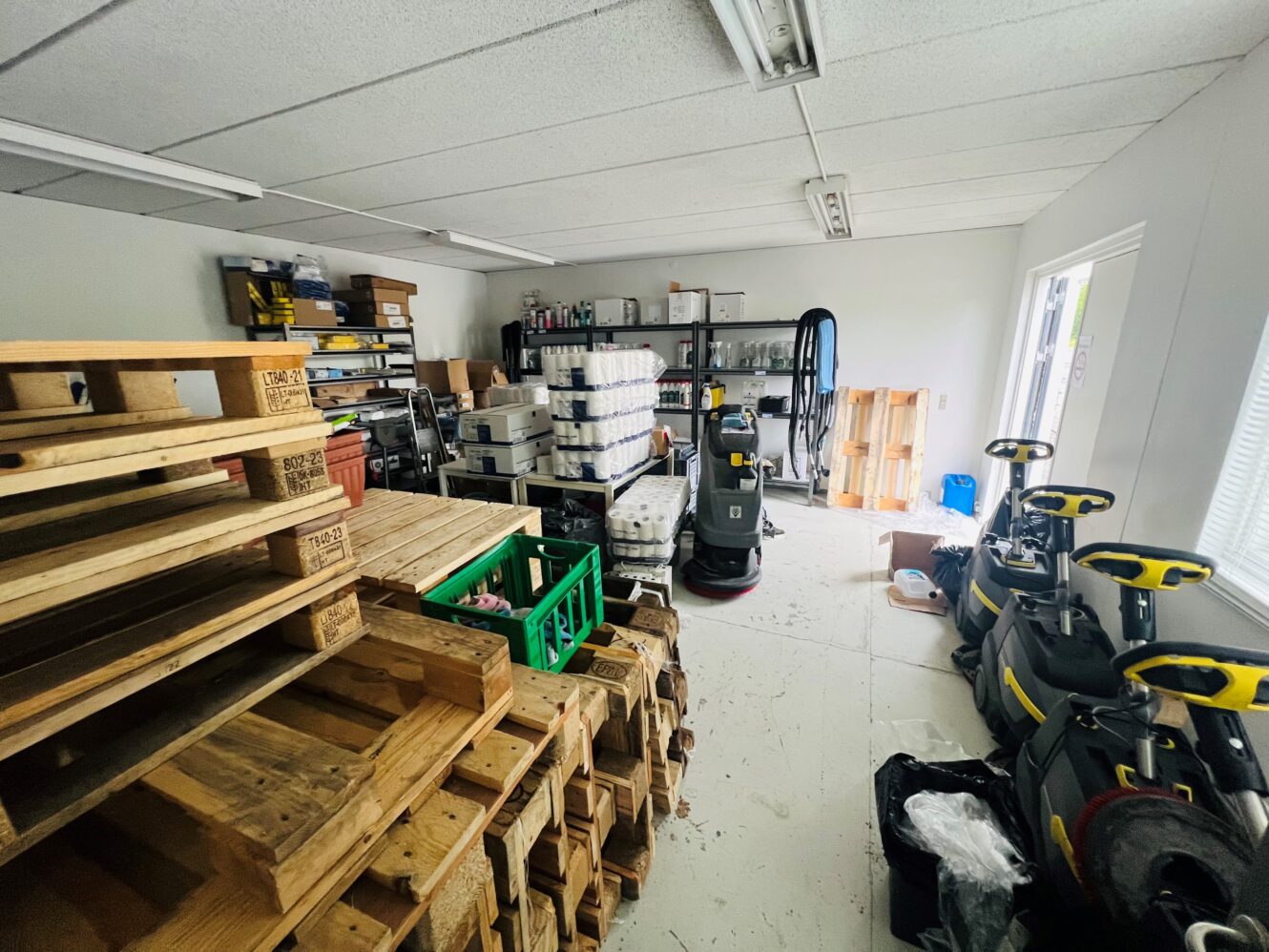 Lagerrum med stablede træpaller, rengøringsmaskiner, hylder med forsyninger og udstyr pænt organiseret langs væggene.