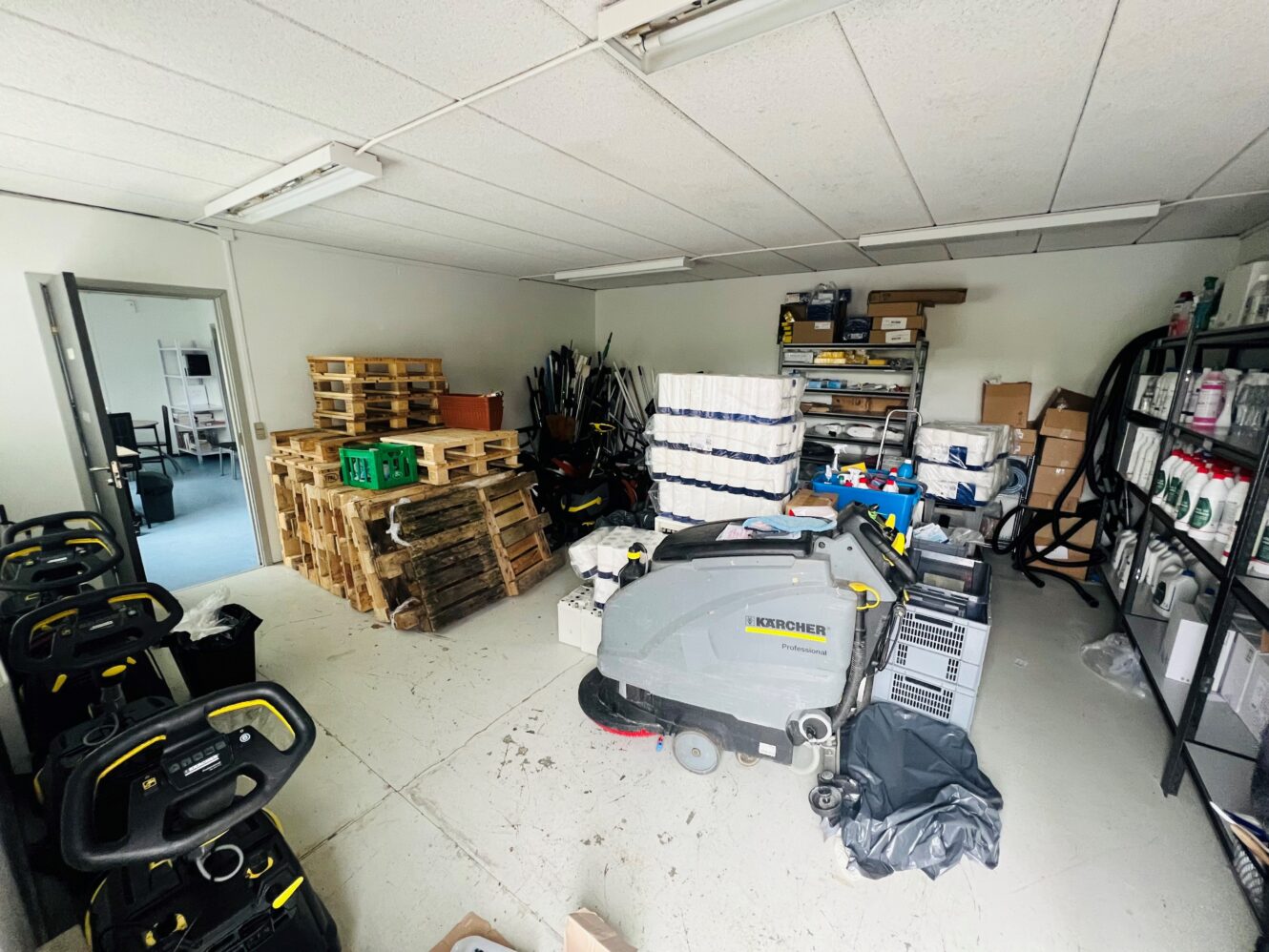 Et lagerrum med rengøringsmaskiner, stablede træpaller, hylder med forsyninger og forskelligt udstyr langs væggene.