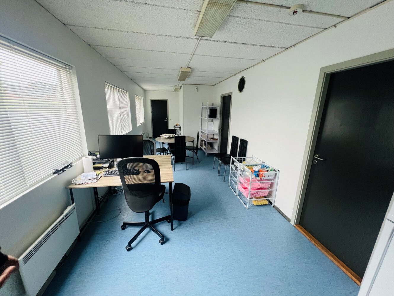 Lille kontorlokale med skriveborde, stole, computere og opbevaringshylder langs hvide vægge. Flere vinduer giver naturligt lys.