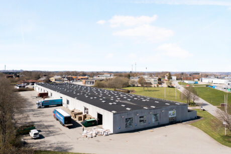 Stort industrilager med læsseramper og lastbiler parkeret udenfor, omgivet af græs, træer og andre industribygninger på en klar dag.