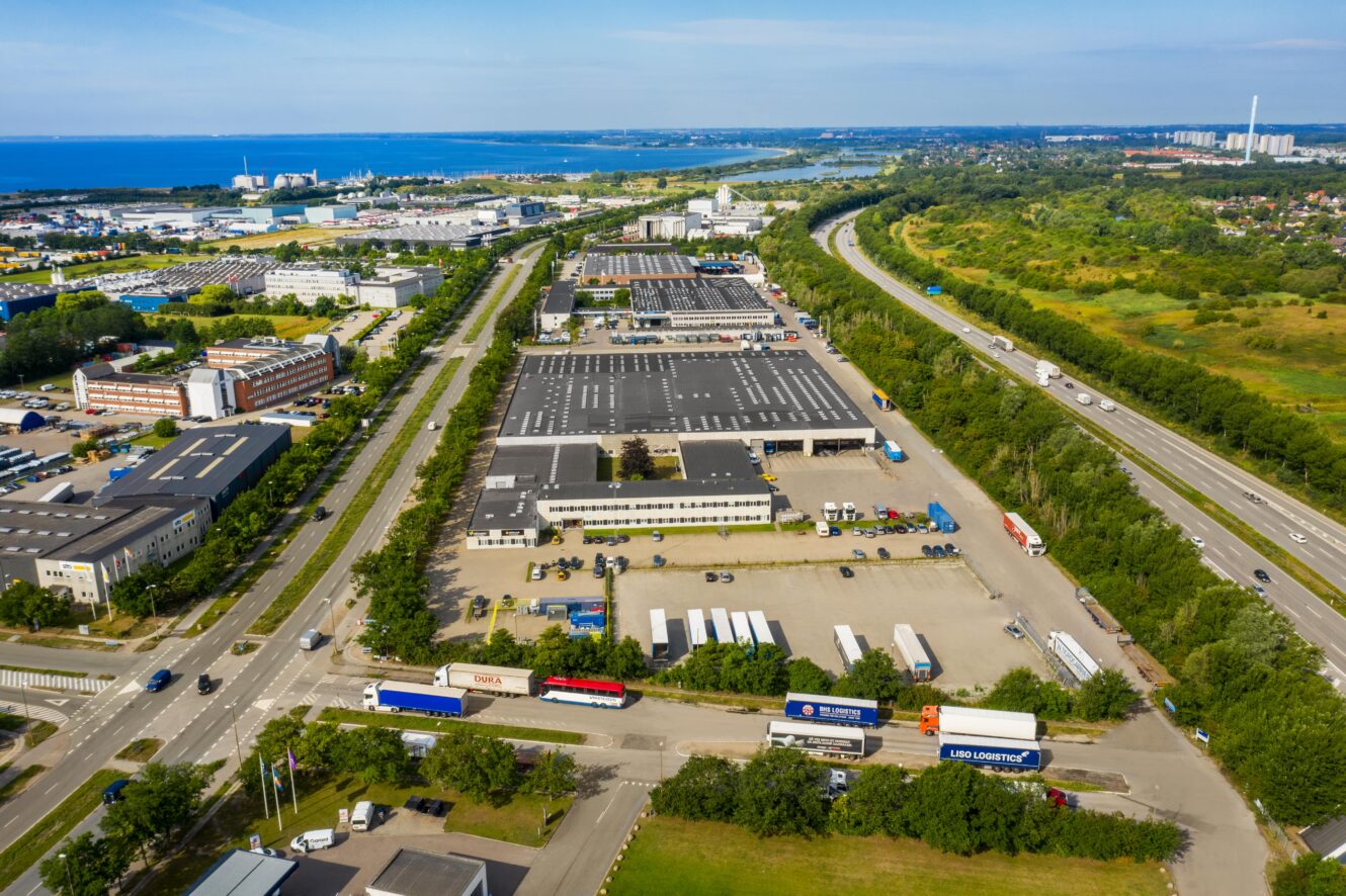 Luftfoto af en industripark med lagerbygninger, lastbiler og nærliggende motorveje, omgivet af grønne områder og fjerntliggende vand i horisonten.