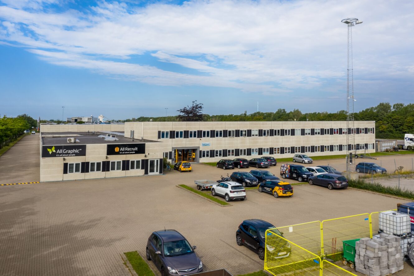 En moderne kontorbygning i to etager med parkeringsplads og flere parkerede biler under en delvist overskyet himmel. Skilte på bygningen viser All Graphic og All Rent.